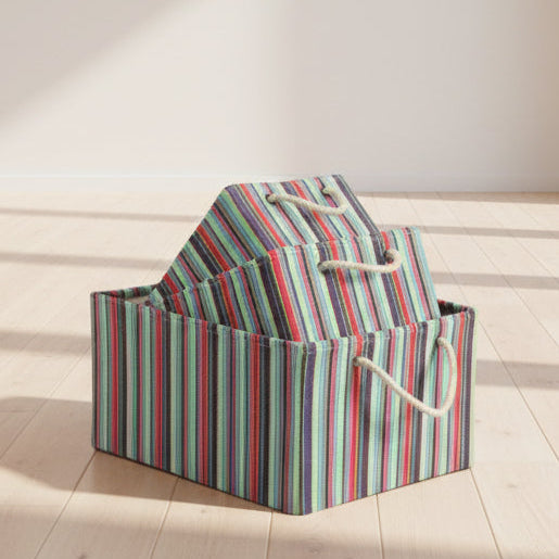 A stack of three fabric storage bins with a colorful vertical stripe pattern in red, green, blue, and gray, featuring cream-colored rope handles, set against a solid off-white background.