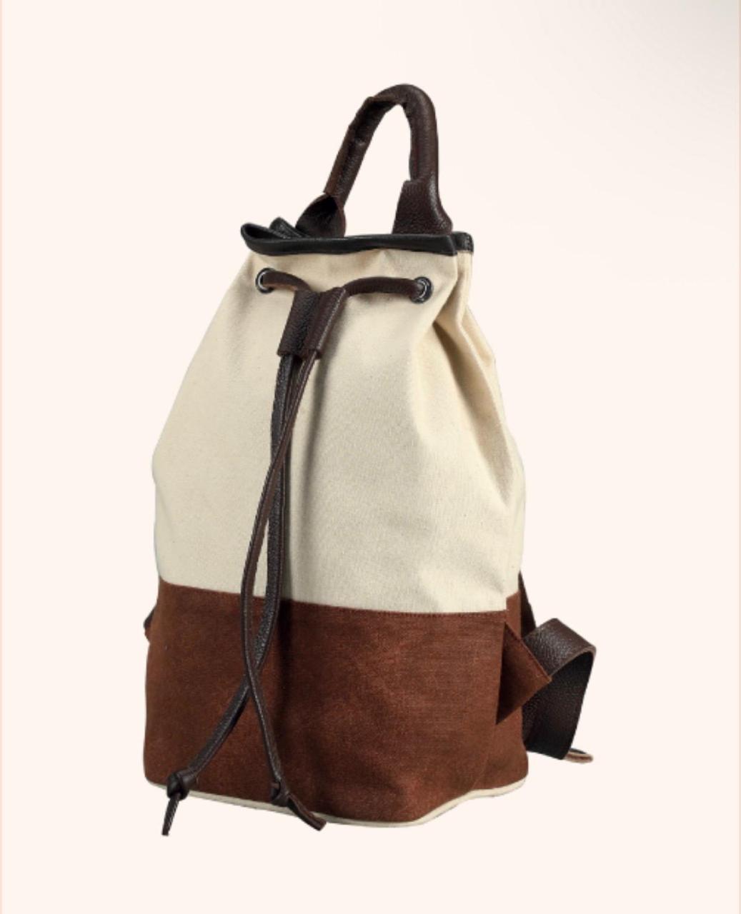 A two-tone canvas backpack with an off-white upper section and a brown lower section, featuring dark brown leather handles, drawstring closure, and side strap on a light cream background.