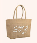 A natural jute tote bag with white embroidered text reading 'SONRI' on the front, set against a solid pale pink background.