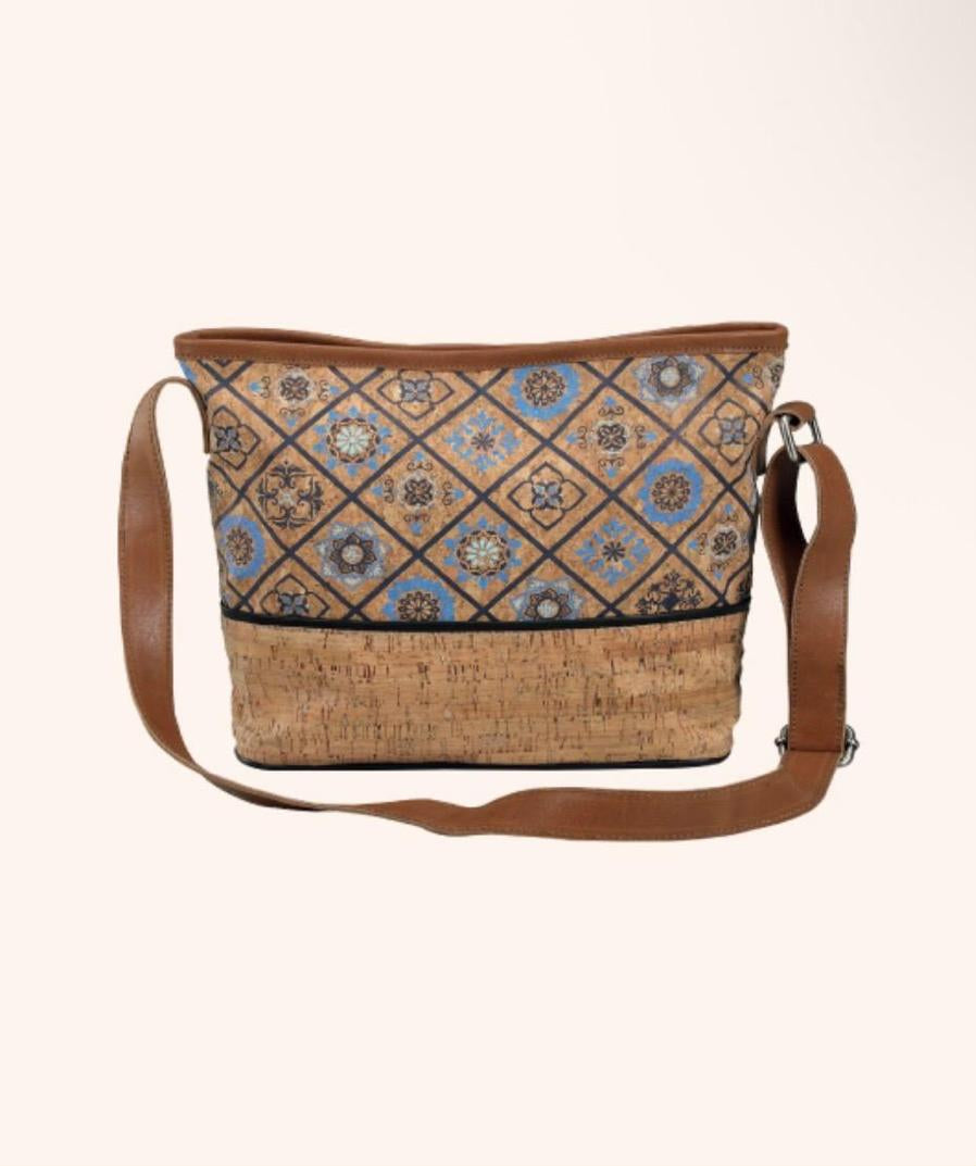 A crossbody bag with a brown leather strap, featuring a patterned upper section with geometric and floral motifs in blue, beige, and brown, and a natural cork-textured lower section.